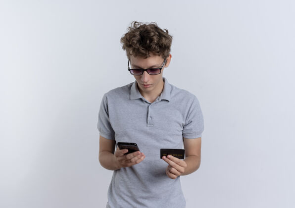 马球一个穿着灰色马球衫的年轻人站在白色的墙上 手里拿着信用卡 看着他的智能手机屏幕屏幕举行年轻