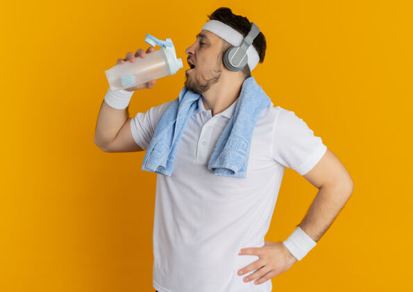 头带身穿白衬衫 头戴头巾 脖子上围着毛巾 站在橙色背景上锻炼后拿着一瓶饮用水的年轻健身男子周围水健身