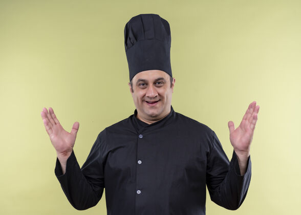 戴上男厨师身穿黑色制服 头戴厨师帽 高举双臂站在绿色背景上 愉快而积极地看着镜头站立帽子举起