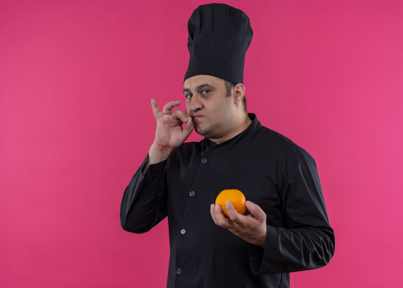 标志男厨师身穿黑色制服 头戴厨师帽 手拿橙色水果 站在粉色背景上展示美味标志立场水果制服