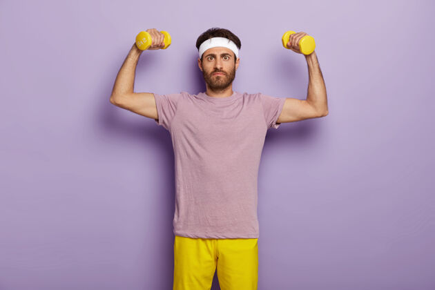 健康运动 锻炼和激励理念严肃的未刮胡子男人举着哑铃举起双臂 穿着紫色t恤和黄色短裤 想要健康强壮运动员个人肌肉