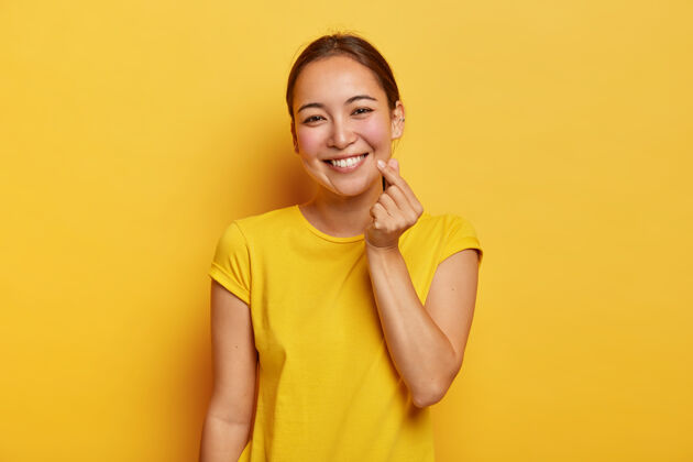 欢呼亚洲外形的快乐女士做韩国人一样的招牌 穿着休闲的黄色t恤有友好的表情站在室内单色镜头肢体语言女性用手势表达爱手穿洞符号