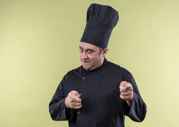 看男厨师身穿黑色制服 头戴厨师帽 看着摄像机 微笑着 用食指指着镜头 镜头站在绿色背景上黑色指向站