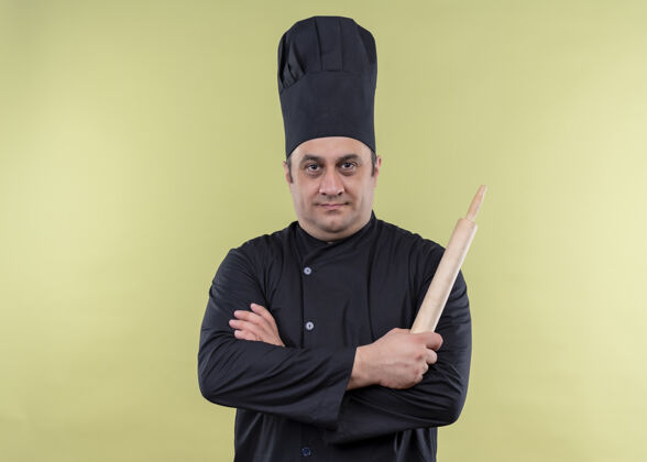 抱着男厨师身穿黑色制服 头戴厨师帽 手持擀面杖 双臂交叉 站在绿色背景上 看着摄像机滚帽子看