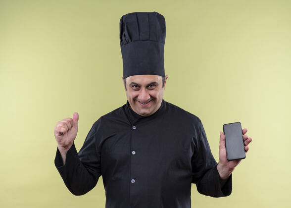 移动男厨师身穿黑色制服 头戴厨师帽 手持智能手机握紧拳头 站在绿色背景上兴奋而快乐制服厨师穿