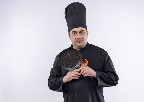 白的男厨师长身穿黑色制服 头戴厨师帽 手持平底锅和木勺 困惑地看着镜头 非常焦急地站在白色背景下煮的木头平底锅