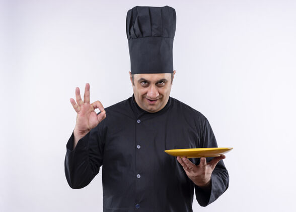 表演男厨师身穿黑色制服 头戴厨师帽 手持盘子 微笑着 白色背景上立着ok标志站着黑色白色