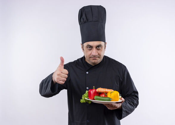 穿男厨师身穿黑色制服 头戴厨师帽 手持新鲜蔬菜的盘子 站在白色背景上竖起大拇指新鲜男盘子