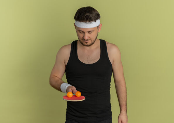 严肃戴着头巾拿着球拍和乒乓球的年轻健身男子站在橄榄色背景下 脸上表情严肃橄榄表情男人