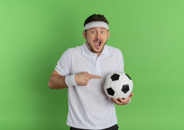 衬衫身穿白衬衫 头箍着足球 食指指着足球的年轻健身男子站在绿色背景上快乐而兴奋站着男士手指