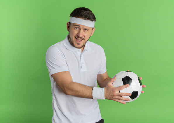 绿色身穿白衬衫 头戴足球带的年轻健身男子站在绿色背景下 兴高采烈地微笑着看着摄像机男士头带足球