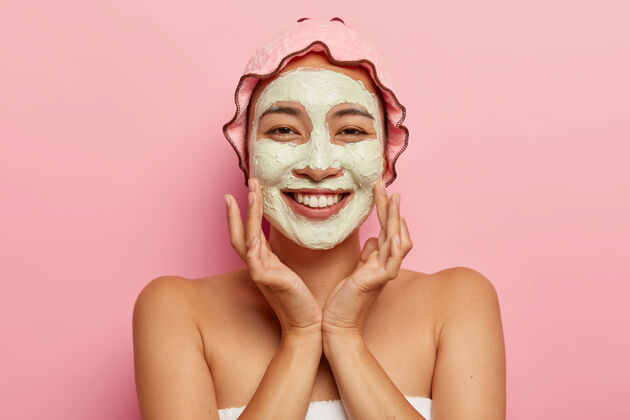 美容师适合所有年龄段的皮肤护理快乐的亚洲女士 脸上涂着去皮泥面膜 做美容 看起来很愉快 摸脸颊 戴着浴帽快乐按摩面膜