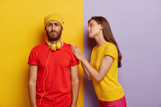 情侣愁眉苦脸的男人有麻烦 戴着黄色的帽子和红色的t恤 不高兴地笑着 关心的女友抚摸着他的肩膀 试图平静和帮助在困难的情况下不满男性人