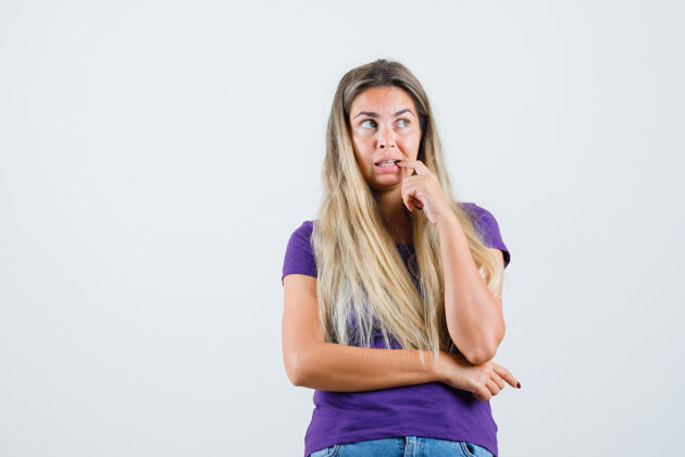 紫罗兰年轻的女士咬着手指在紫罗兰色t恤 牛仔裤和看起来焦虑 前视图人咬黑发