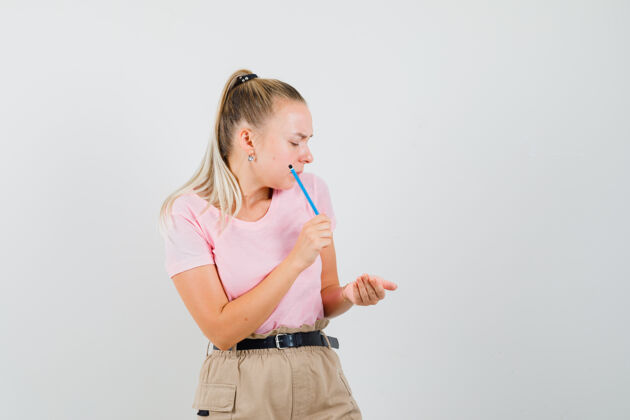 年轻年轻的女性拿着铅笔 看着手捧着杯状t恤 裤子的前视图成人人美丽