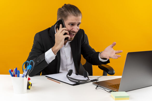 英俊沮丧的帅哥商人穿着西装在笔记本电脑上工作 在手机上聊天 在橙色背景下坐在办公桌旁 神情困惑和不快交谈电话西装