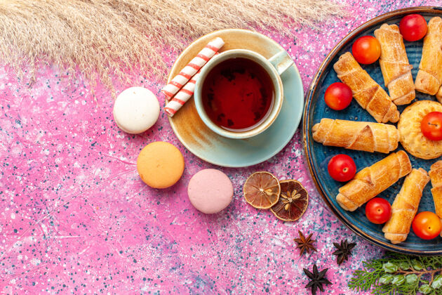 李子顶视图甜甜可口的百吉饼在托盘内与新鲜酸李子法国马卡龙和一杯茶在淡粉色的桌子上法式蛋糕食物