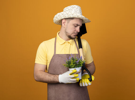 花园年轻的园丁穿着连体衣 戴着帽子 拿着铲子和盆栽植物 站在橘色的墙上 带着悲伤的表情往下看表情人罐子