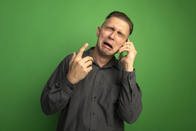 灰色一个穿着灰色衬衫的年轻帅哥站在绿色的墙上一边打电话一边哭年轻手机人