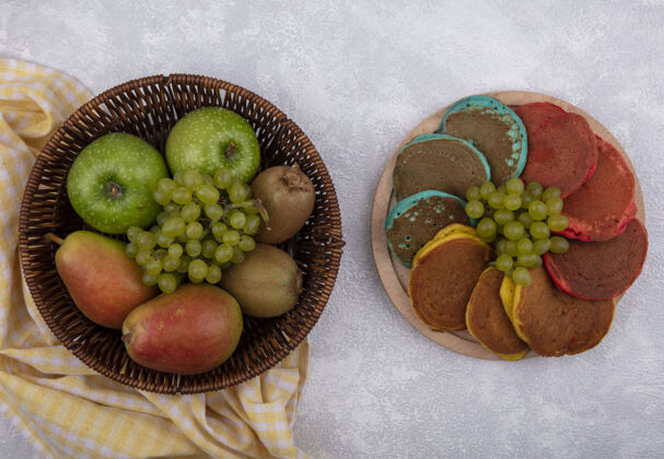 葡萄俯瞰图：黄色格子毛巾上的篮子里放着梨子 青苹果 葡萄和猕猴桃 白色背景上的架子上放着彩色的煎饼食物颜色白色