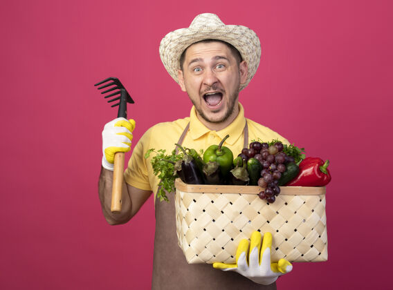 蔬菜年轻的园丁穿着连体衣 戴着帽子 戴着工作手套 手里拿着装满蔬菜的箱子 手里拿着小耙子 站在粉色的墙上兴奋而快乐地喊道耙子胡椒穿