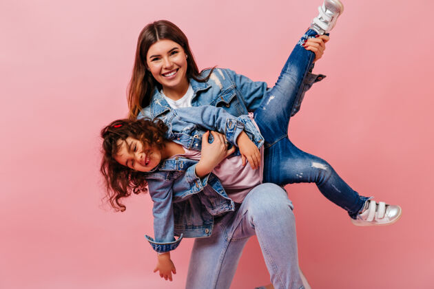 成人迷人的女人和小女儿在粉色背景下玩耍妈妈和未成年的孩子穿着牛仔夹克的摄影棚照片黑发幸福为人父母