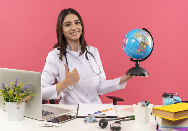 看年轻的女医生穿着白大褂 脖子上戴着听诊器 拿着地球仪 面带微笑地看着前方 竖起大拇指坐在桌旁 笔记本电脑放在粉红色的墙上医院女性办公室