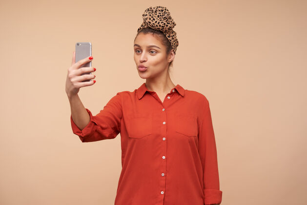 漂亮一位面容讨人喜欢的棕色头发的年轻女子 穿着休闲装站在米色的墙上 一边用手机拍着自己的照片 一边噘着嘴唇姿势穿模特