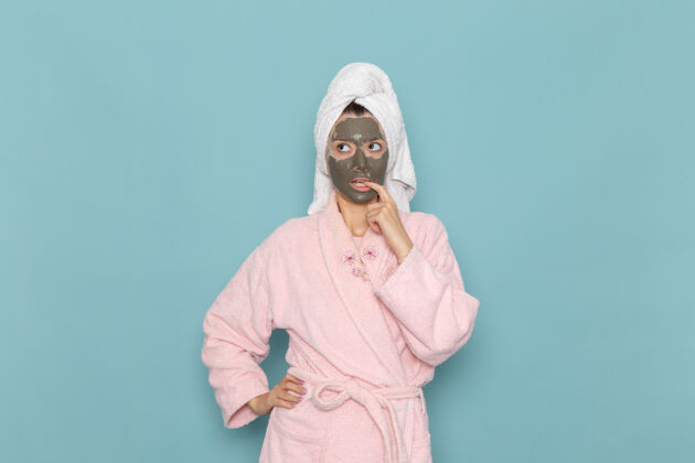 面具正面图身着粉色浴袍 脸上戴着面具的年轻女性思考蓝色墙壁淋浴清洁美容自护霜年轻人面部淋浴