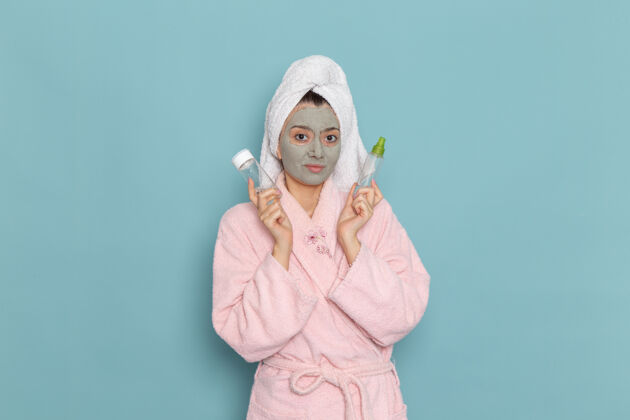 前面正面图身穿粉色浴袍的年轻女性 脸上戴着口罩 拿着蓝色墙上的喷雾剂淋浴清洁美容自护霜自我护理面部年轻人