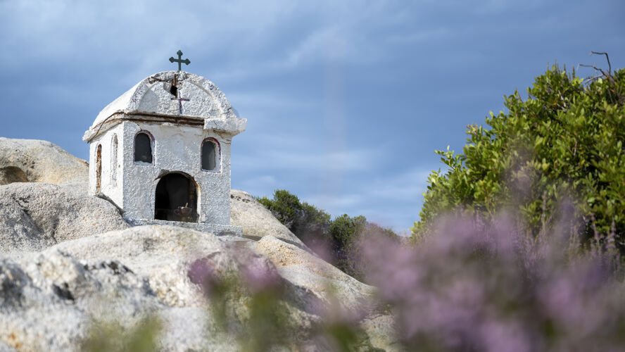 东正教一座古老而小巧的神龛坐落在爱琴海沿岸的岩石上 四周灌木丛生 天空多云 希腊纪念碑教十字架微型