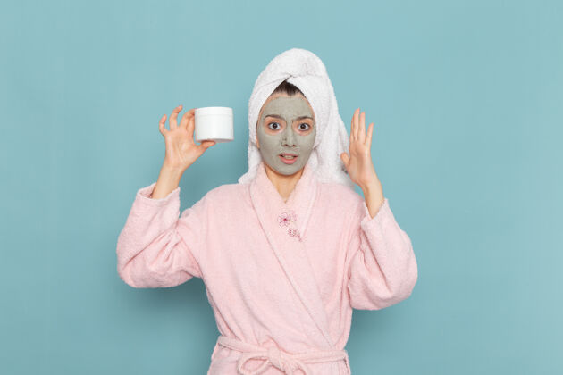 视图正面图身穿粉色浴袍的年轻女性 脸上戴着口罩 蓝色墙上拿着面霜淋浴 清洁美容自我护理面霜人女性清洁