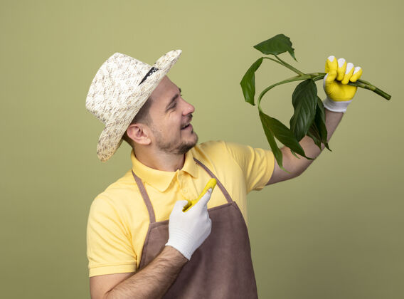 年轻人年轻的园丁 穿着连体衣 戴着帽子 戴着工作手套 手里拿着一株植物 用食指指着它 站在明亮的墙壁上 兴高采烈地笑着光制服举行