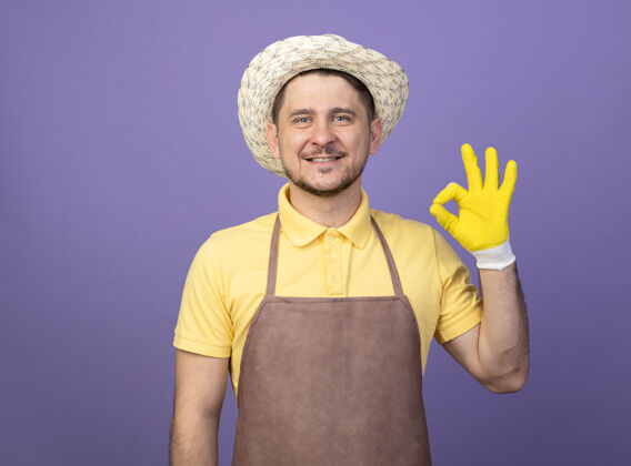 制服年轻的园丁身穿连体衣 戴着工作手套 戴着帽子 面带微笑 站在紫色的墙上 脸上挂着“ok”的牌子标志微笑花园