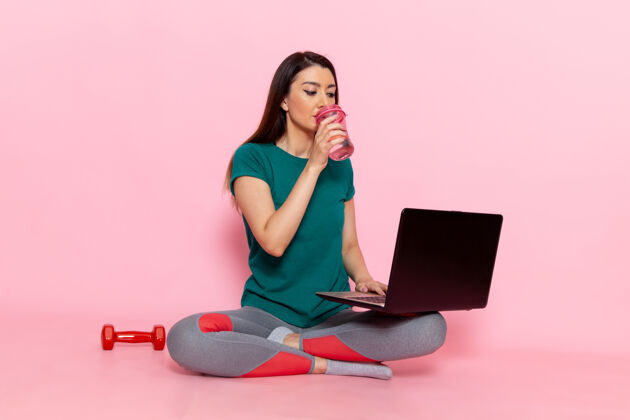 运动前视图身着绿色t恤的年轻女性在浅粉色墙壁上使用笔记本电脑腰部运动锻炼美丽苗条的女运动员前运动腰
