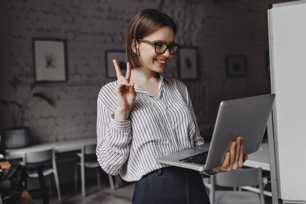 技术戴着耳机和眼镜的积极女孩展示和平标志 在工作场所的笔记本电脑上用视频交谈肖像工作场所通信