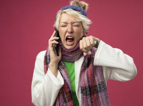 电话愤怒 沮丧 生病 不健康的女人 短发 戴着暖和的围巾和帽子 站在粉红色的墙上一边打电话一边感觉不舒服围巾喊叫不舒服