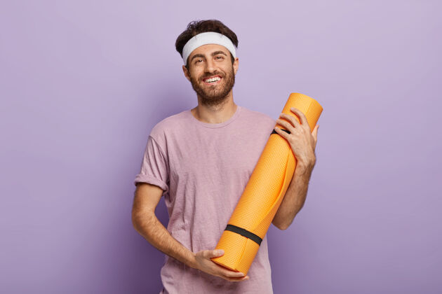 小伙子高兴的剃须男拿着卷起的健身垫 满意的瑜伽课后衣服运动教练