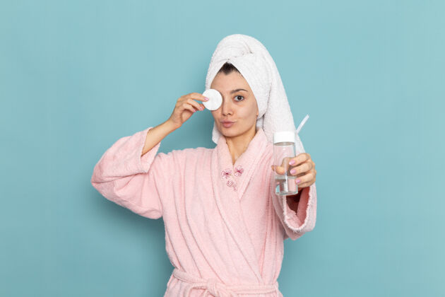 年轻人正面图身着粉色浴袍的年轻女性在蓝色墙壁上洗脸化妆淋浴清洁美容自护霜浴袍清洁奶油