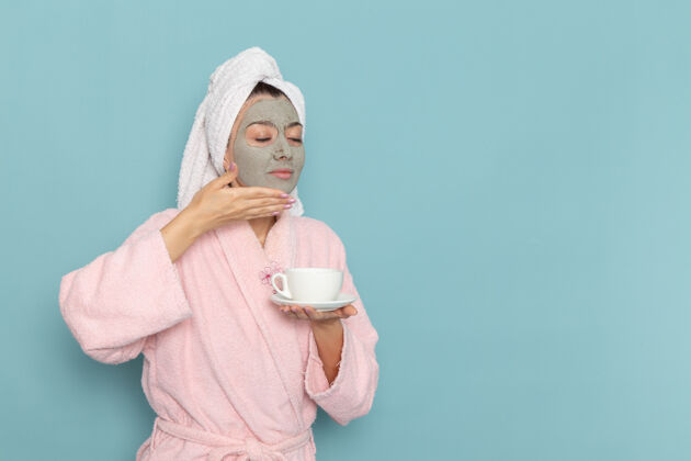 女性正面图身穿粉色浴袍 脸上戴着口罩的年轻女性拿着咖啡在蓝色墙上淋浴清洗美容自护霜咖啡保持面霜