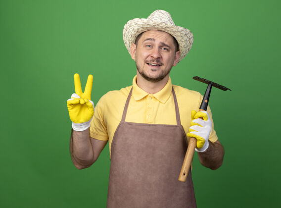 手套年轻的园丁穿着连体衣 戴着帽子 戴着工作手套 拿着小耙子 看着前面的v形标志 微笑着站在绿色的墙上微笑人人