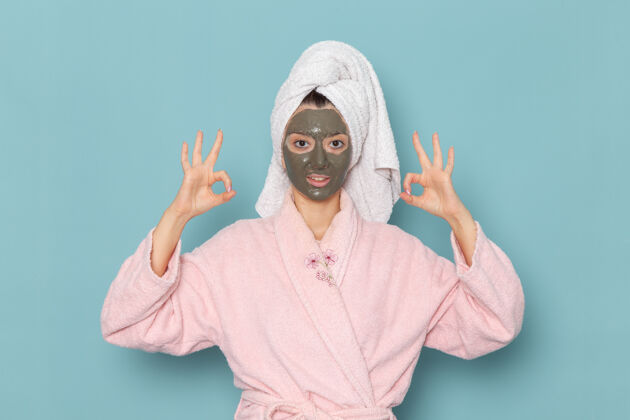 视野正面图身着粉色浴袍的年轻女性在蓝色墙壁上戴着面具摆姿势美丽的水浴霜自我护理淋浴女性自我照顾淋浴