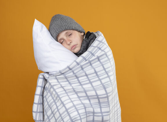 抱着生病的小男孩戴着暖和的帽子 裹着毛毯发烧 抱着枕头站在橙色的墙上毯子帽子包裹