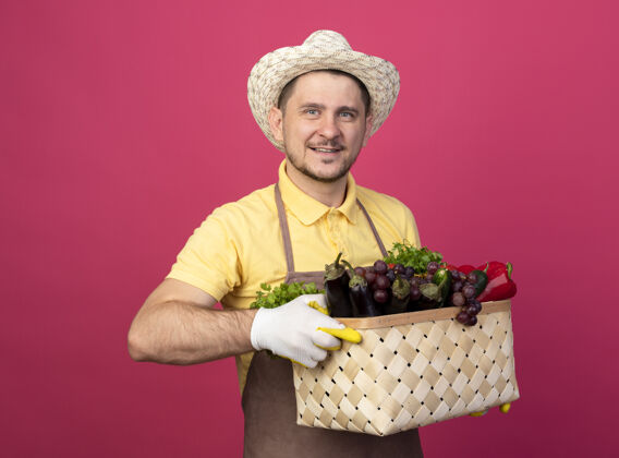 人年轻的园丁穿着连体衣 戴着帽子 戴着工作手套 手里拿着装满蔬菜的箱子 面带微笑地看着前面 站在粉色的墙上 快乐而积极工作花园粉色