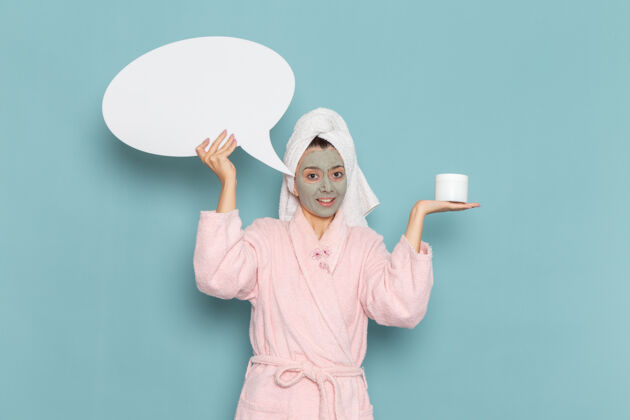 人正面图身穿粉色浴袍的年轻女性 脸上戴着口罩拿着面霜 蓝色墙壁上有白色标志淋浴清洁美容自护霜自我护理前面人