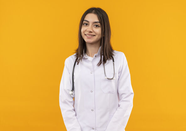 姿势身穿白大褂 脖子上戴着听诊器的年轻女医生站在橘色的墙上 带着自信的微笑望着前方医生看自信