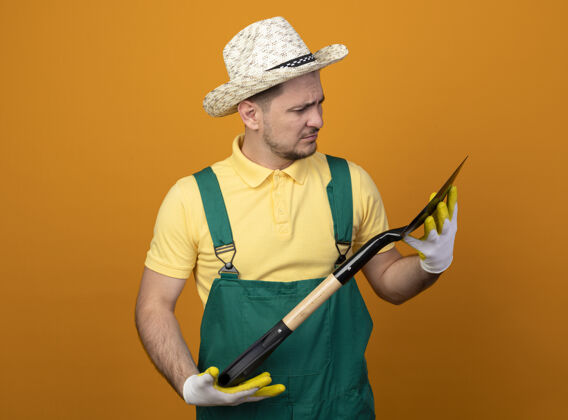 市民年轻的园丁穿着连体衣 拿着铲子戴着帽子 站在橙色的墙上严肃地看着它人工人橙色