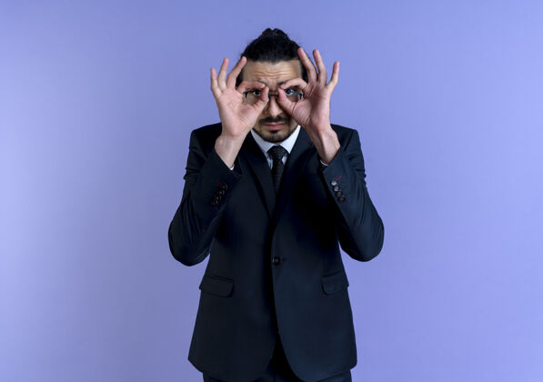 人一个穿着黑西装戴着眼镜的商人站在蓝色的墙上做着双眼手势 手指透过手指看公司手指西装