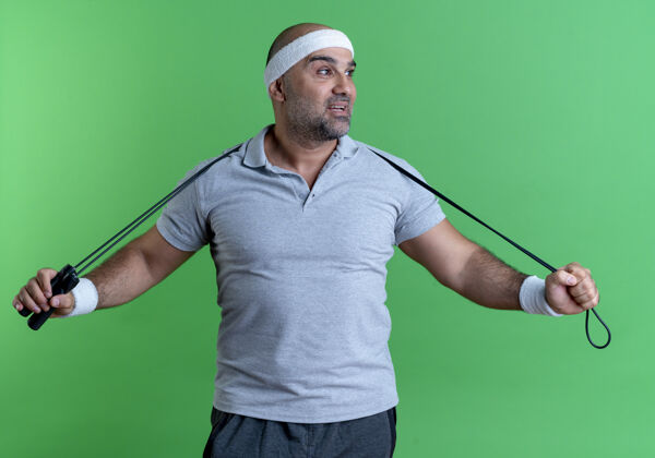 绳子戴着头巾 手拿跳绳 脸上带着微笑的成熟运动型男人站在绿色的墙上人抱着人