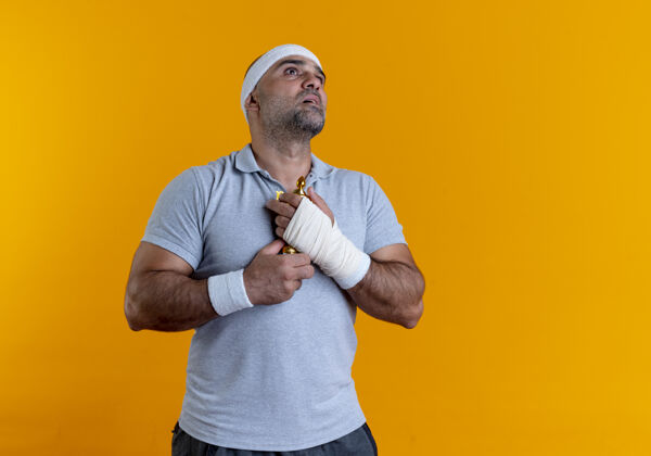教练戴着头巾的成熟运动型男人抱着他的奖杯 站在橙色的墙上 带着希望的表情仰望着运动装运动人